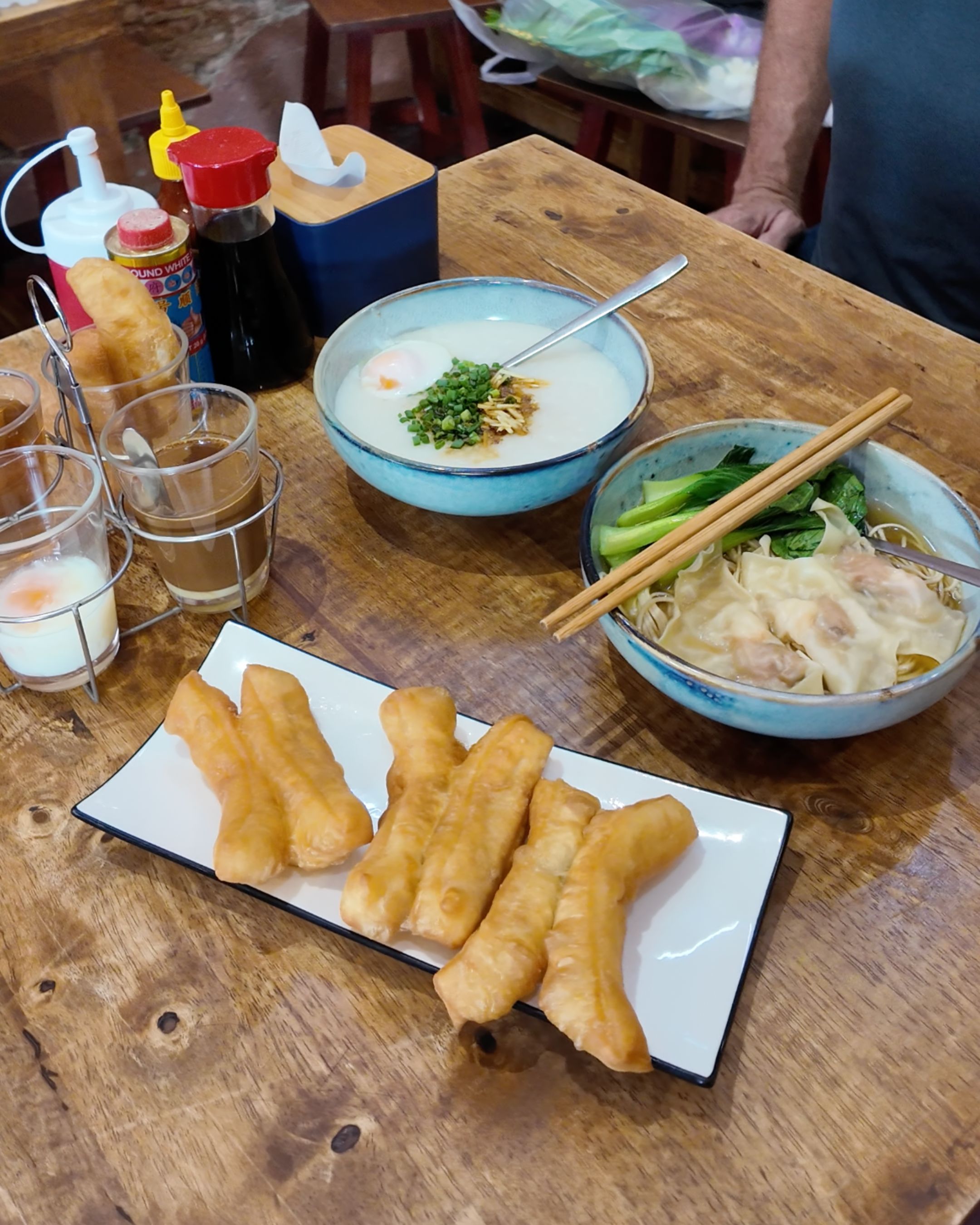 A selection of traditional Thai-Chinese breakfast dishes, including rice porridge, wonton noodles and crispy fried dough sticks served together.