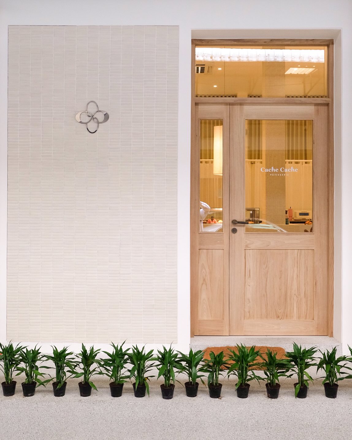 Modern restaurant exterior at dusk with warm indoor lighting visible through large glass windows. The entrance is framed by lush green plants and elegant white pillars under a clean, minimalist white facade